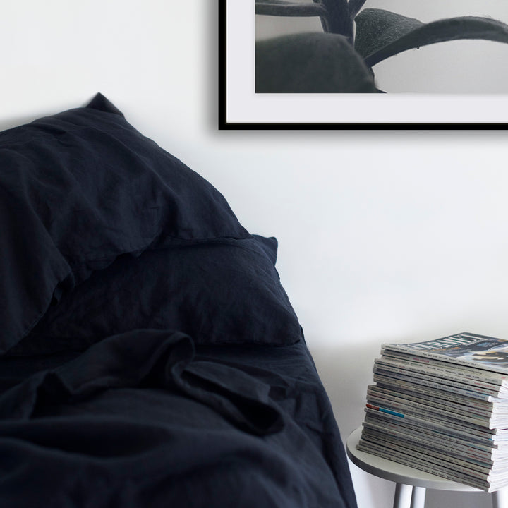 A cropped photo of a bed dressed in Navy bed linen. There is a white bedside table next to the bed with a stack of magazines on top of it. Sizes: Queen, King, Super King