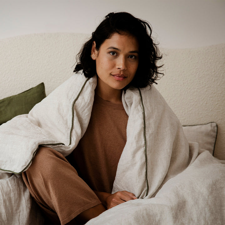 A woman sitting on a bed, with a Natural and Forest Piped linen Duvet Cover over her shoulders. Size: Queen, King