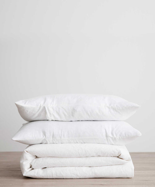Folded white linen duvet cover set with two soft pillows stacked on light wooden surface against plain background