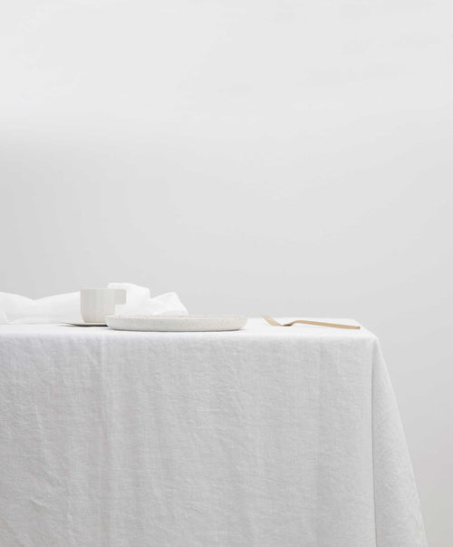 Linen Tablecloth - White draped on a table with simple white dishware and light wooden cutlery The beauty of home