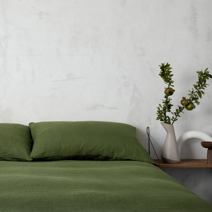 Close up of a bed dressed in Forest bedlinen, styled with a wooden side table and vase with greenery.							