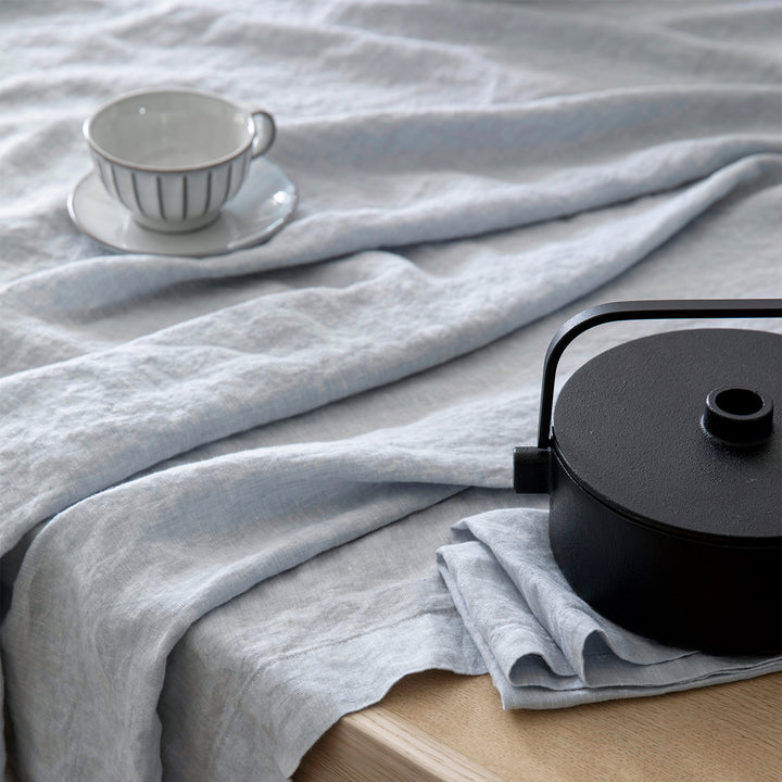 Close up of a table dressed in a Linen Tablecloth in Sky and a Linen Napkin in Sky, styled with a black iron teapot and small white teacup and saucer.