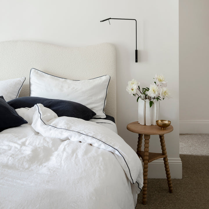 A cream bed dressed in White and Navy Piped bedlinen with a Navy Fitted Sheet and Pillowcases is styled with a side table and vase of flowers.