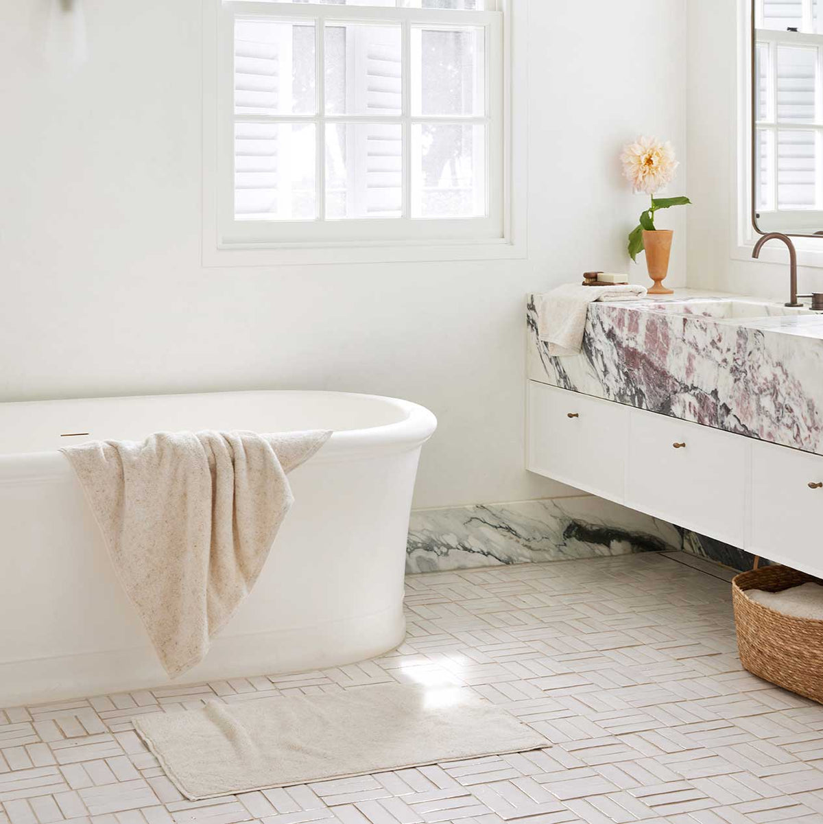 Bright bathroom with white freestanding bath, soft linen towel, marble vanity, and woven basket below sink