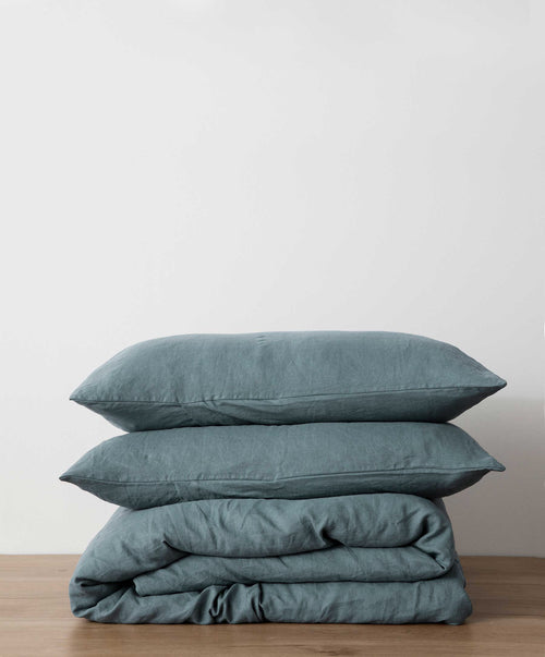 Bluestone linen duvet cover set folded with two matching pillowcases on wooden surface against neutral background
