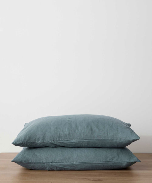 A Set of 2 Linen Pillowcases in Bluestone, made from 100% European flax, are stacked on a wooden surface with a plain white background.