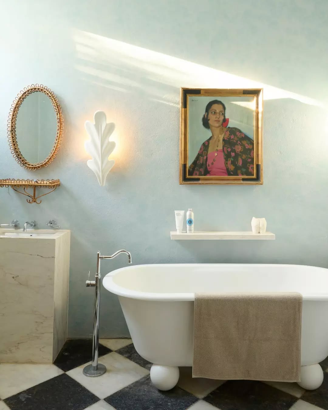 Bathroom with clawfoot tub, beige towel, decorative wall sconce, oval mirror, and framed portrait above tub