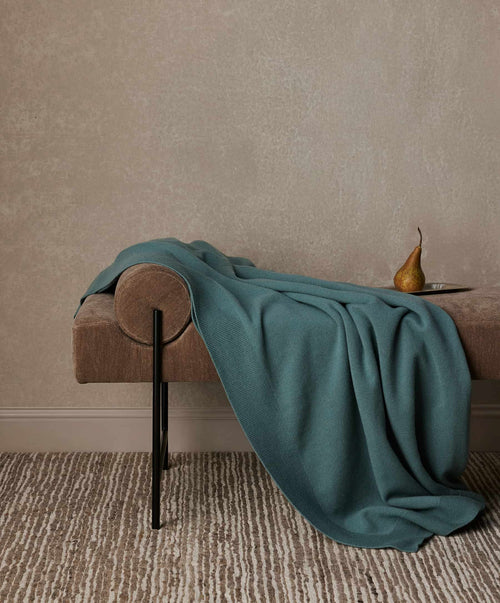 Teal blanket draped over a wooden bench against a beige wall.