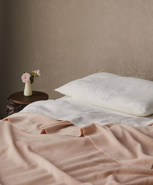 Neatly made bed with pink blanket and white pillows, small table with flowers against a beige wall.