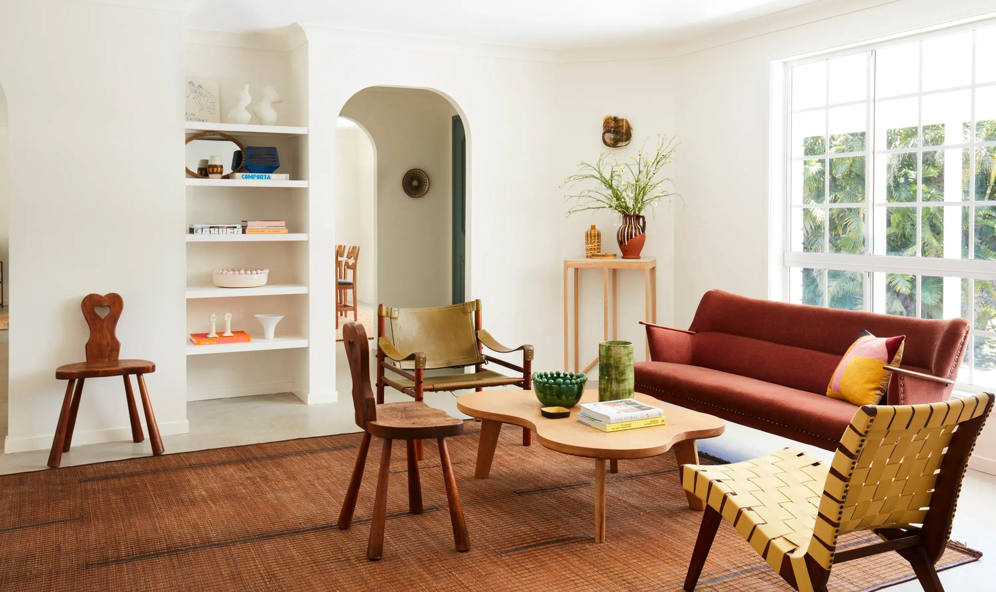A styled living room interior featuring a rust-red sofa, wooden chairs, a woven yellow armchair, a round wooden coffee table, and a large window on a white wall.