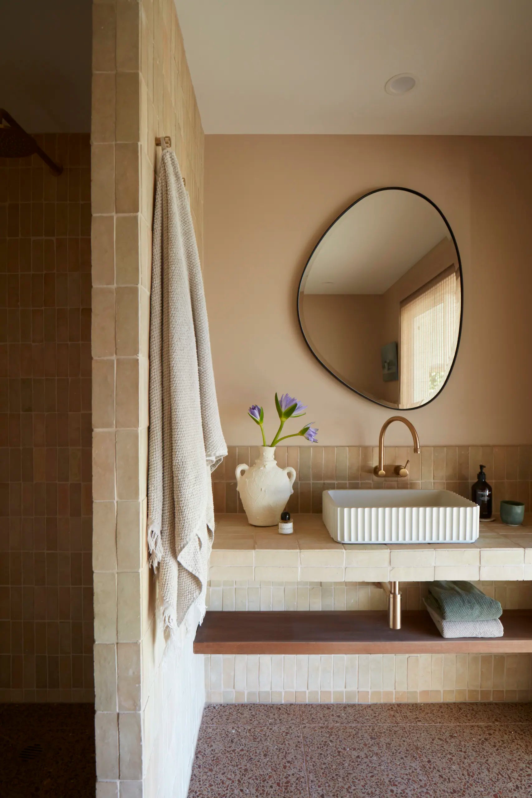 A modern bathroom vanity featuring a rectangular white vessel sink with a ribbed front, a curved brass faucet, a round black-framed mirror, and a beige tiled backsplash. A textured cream towel hangs on the tiled wall to the left, and a small white vase with purple flowers sits on the counter.