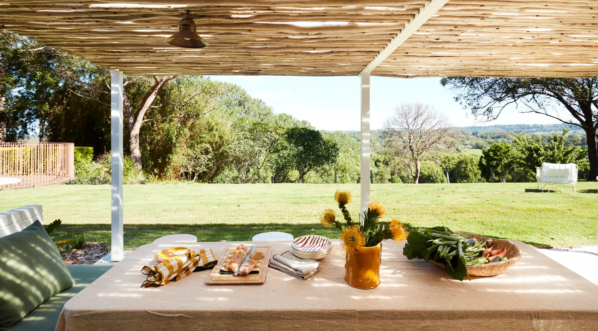 Outdoor dining setup under a thatched roof pergola with a table set for guests, featuring a yellow vase of sunflowers, folded napkins, and a basket of greens, overlooking a lush green lawn and trees.