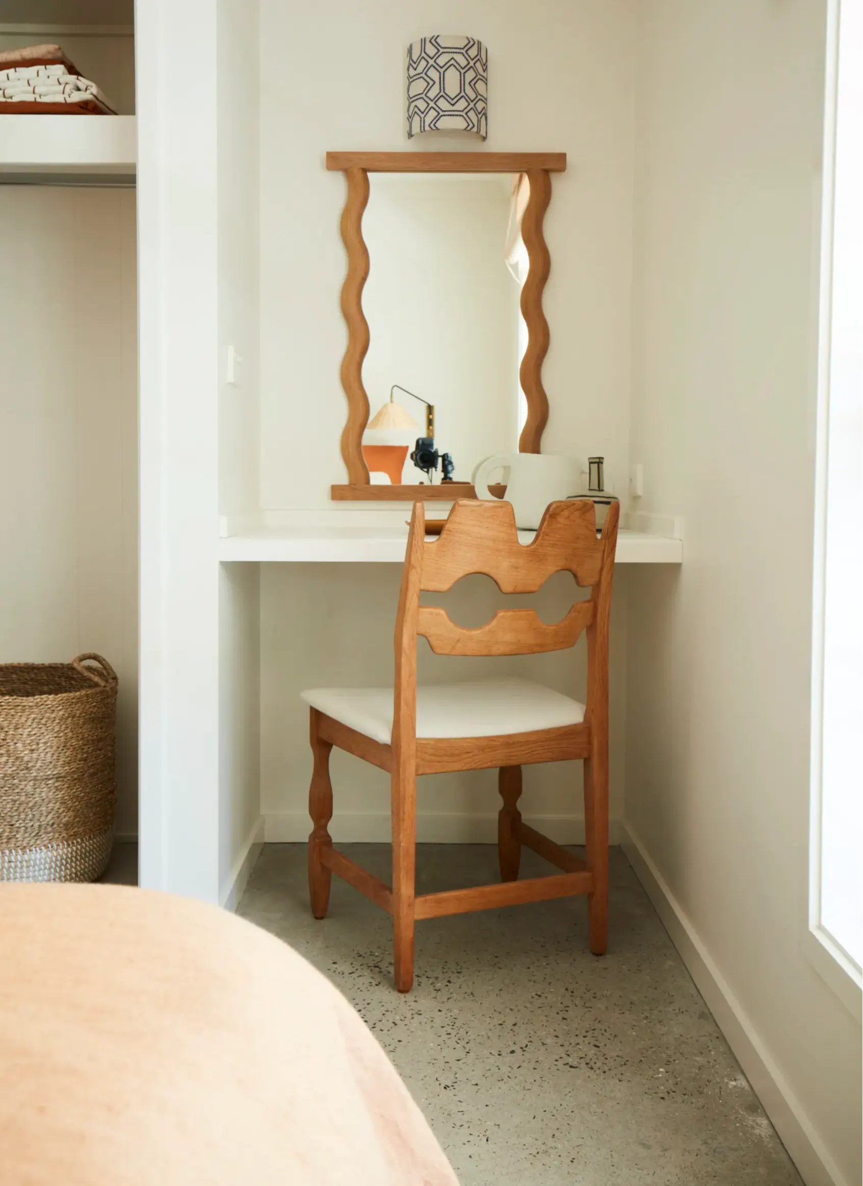 A wooden vanity desk with a matching scalloped mirror and a carved wooden chair with a light cushion, set in a bright, minimalist room.