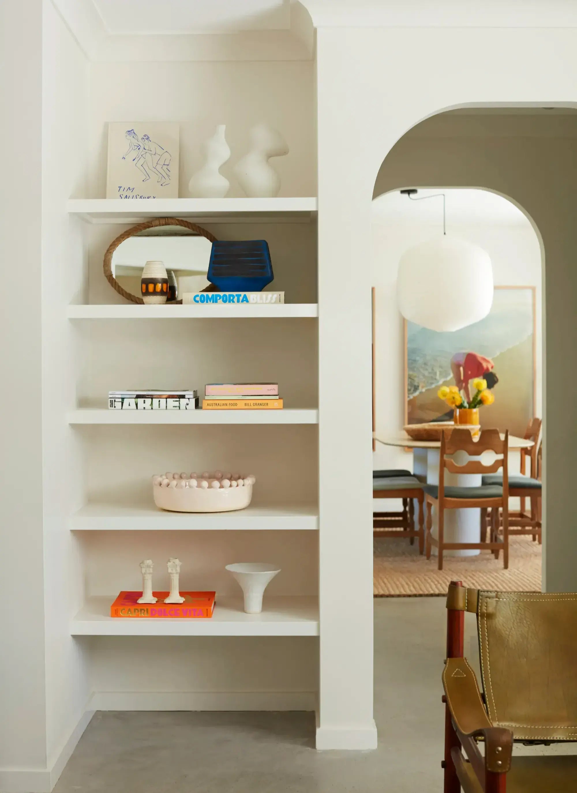 A tall white built-in shelving unit with five shelves styled with books, vases, a small mirror, and decorative objects, set in a bright modern interior with an arched doorway leading to a dining area.