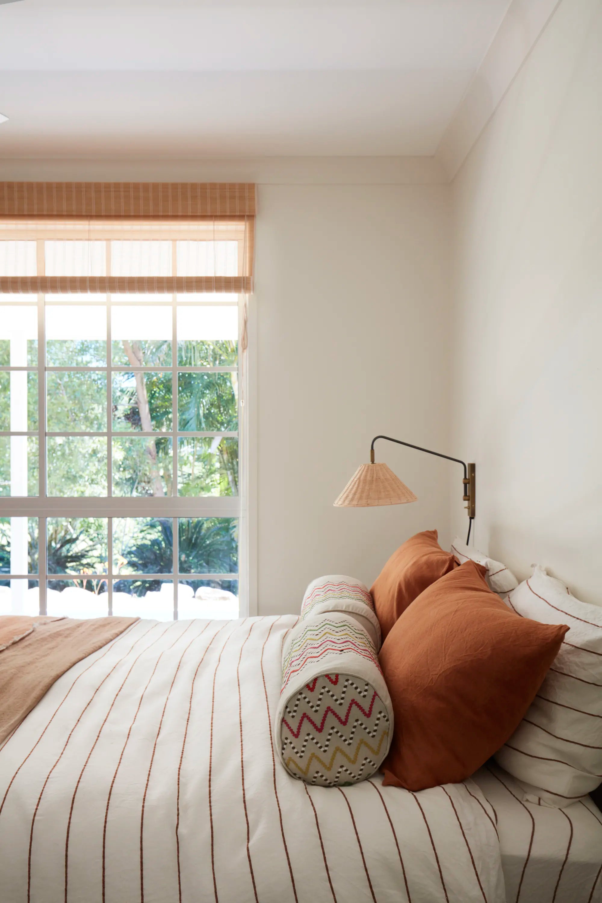 A styled bedroom scene featuring a bed with cream and brown striped bedding, rust-colored accent pillows, a patterned cylindrical bolster pillow, a wall-mounted lamp, and a window with a bamboo roller shade overlooking greenery.
