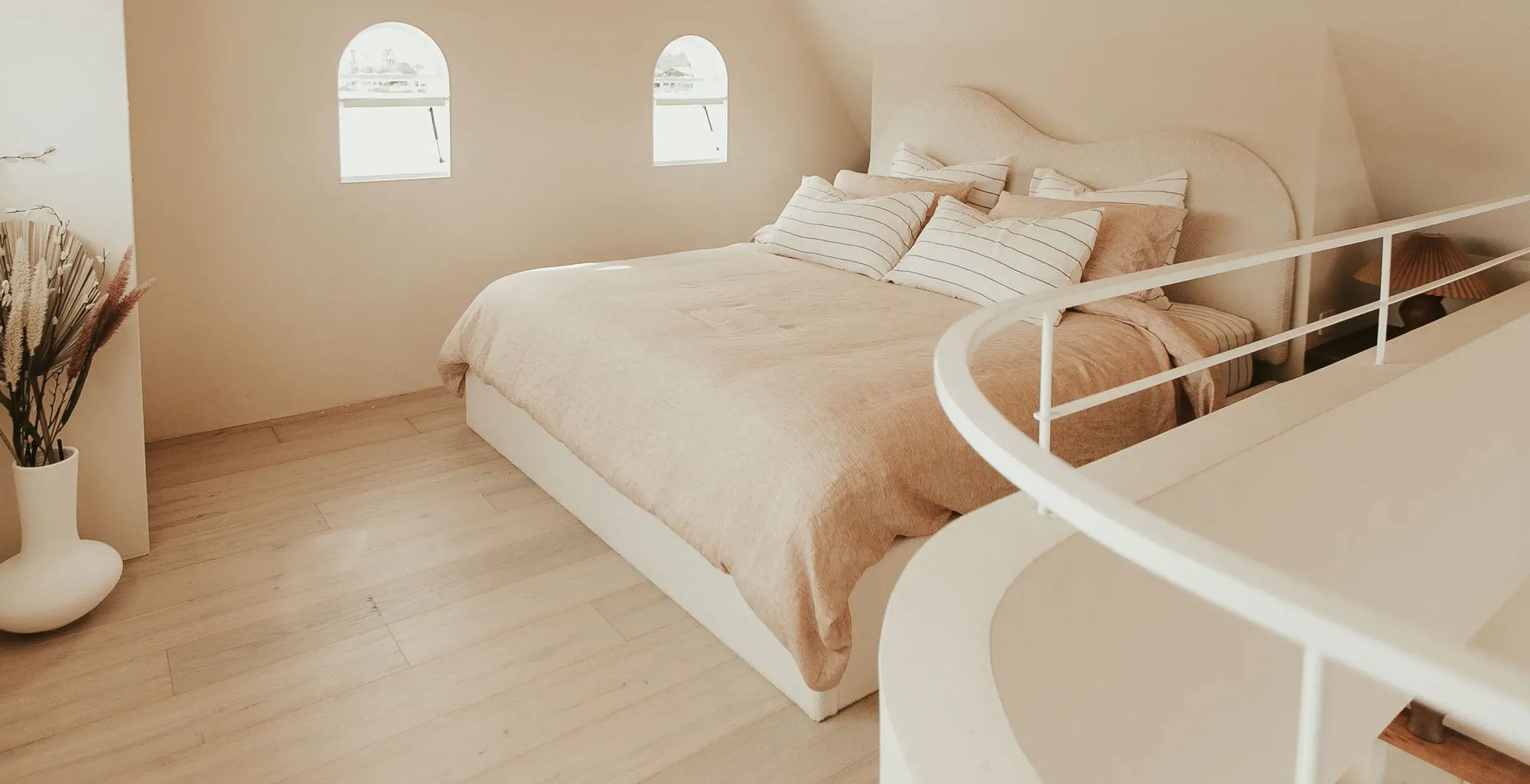 A minimalist bedroom featuring a bed dressed in beige and white linens, set in a warm, cream-colored room with light wood floors and arched windows. A white curved staircase railing is visible in the foreground.