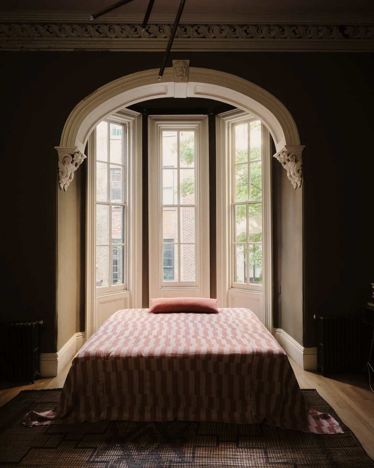 A single bed with a pink and white striped patterned bedspread and a matching pillow, positioned in a dark room beneath a large arched bay window.
