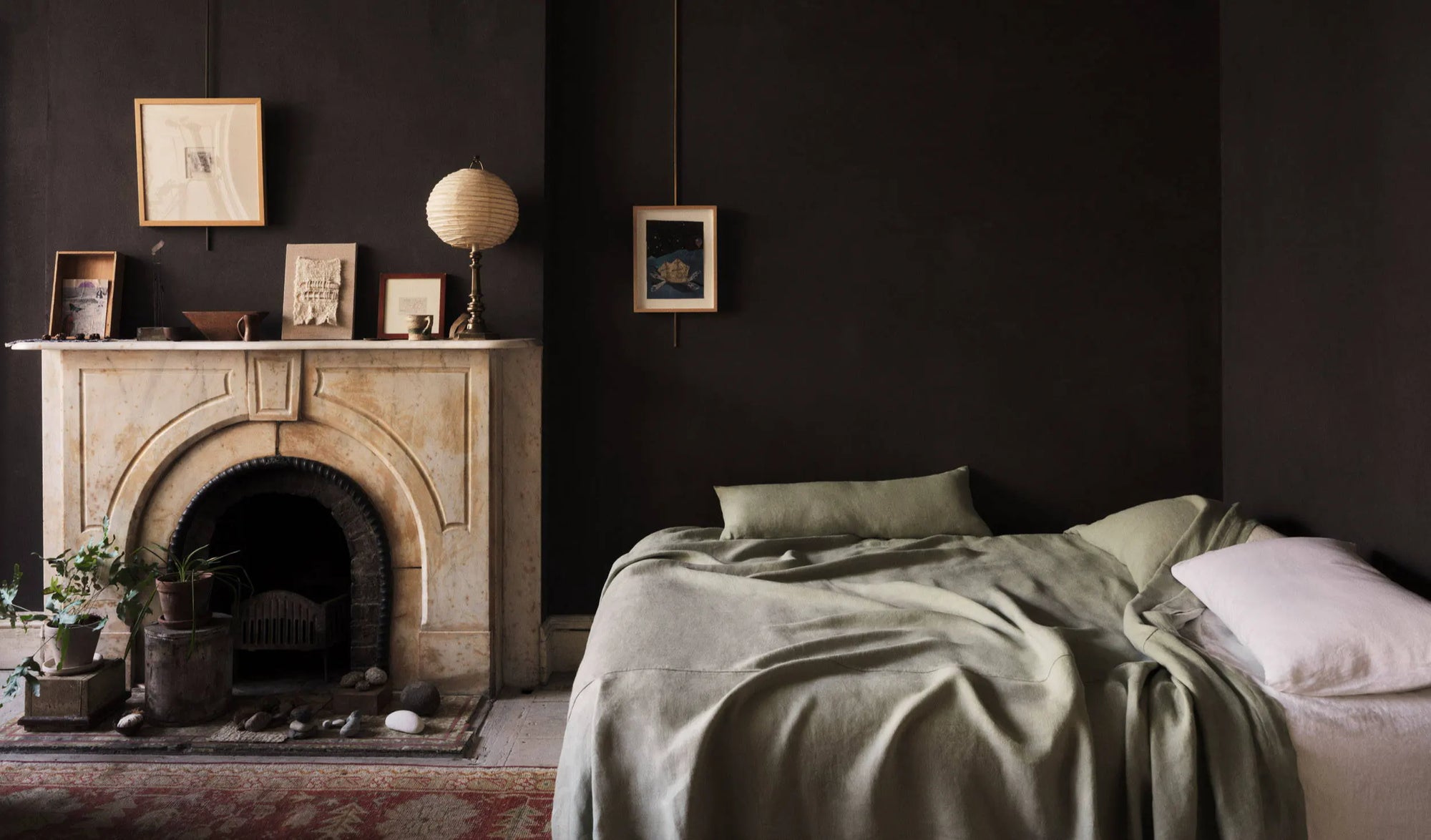 A styled bedroom scene with a bed dressed in muted sage green bedding and white pillows, set against dark charcoal walls beside a stone fireplace with framed art and a lamp.