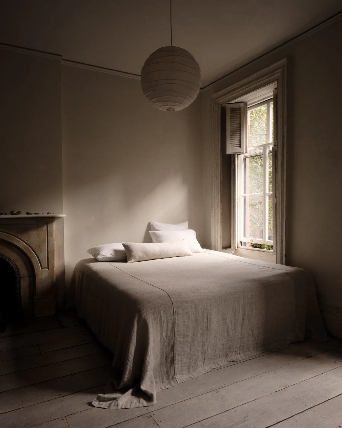 A minimalist bedroom scene featuring a neatly made bed with white linens and pillows, a spherical white paper lantern hanging from the ceiling, a window with shutters, and a stone fireplace on the left, all in soft natural light.