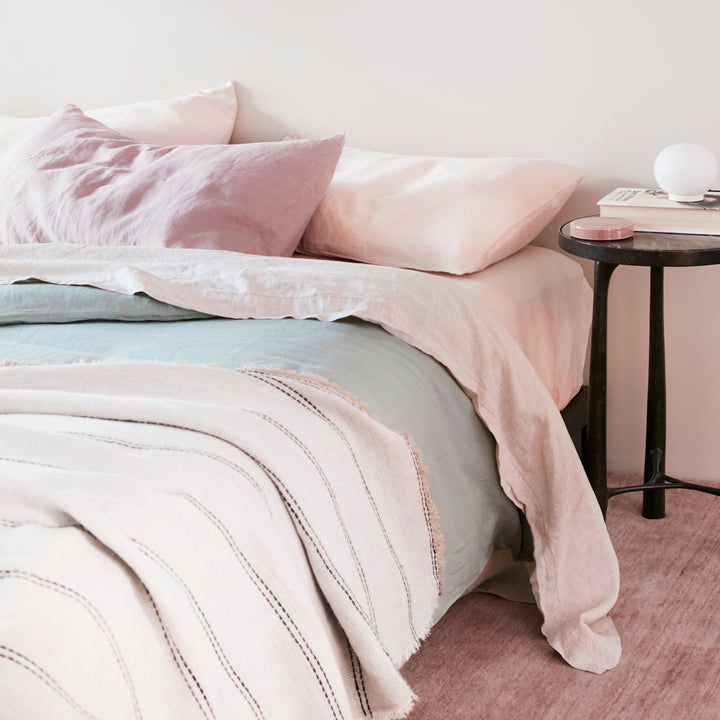 A bed dressed in a Sheet Set in Blush with a Pillowcase in Dusk, a Duvet Cover in Sage and a Mira Ana Throw. A black side table holds a small lamp and a book. Sizes: Queen, King