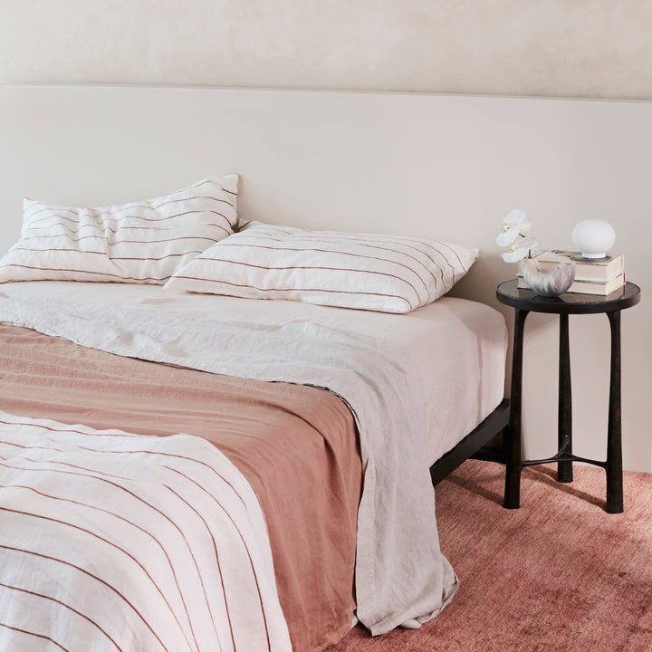 A bed dressed in Cedar Stripe, Fawn, Smoke Grey and Blush bed linen, styled with a black bedside table, small vase and a couple of books. Sizes: Single, Double, Queen, King