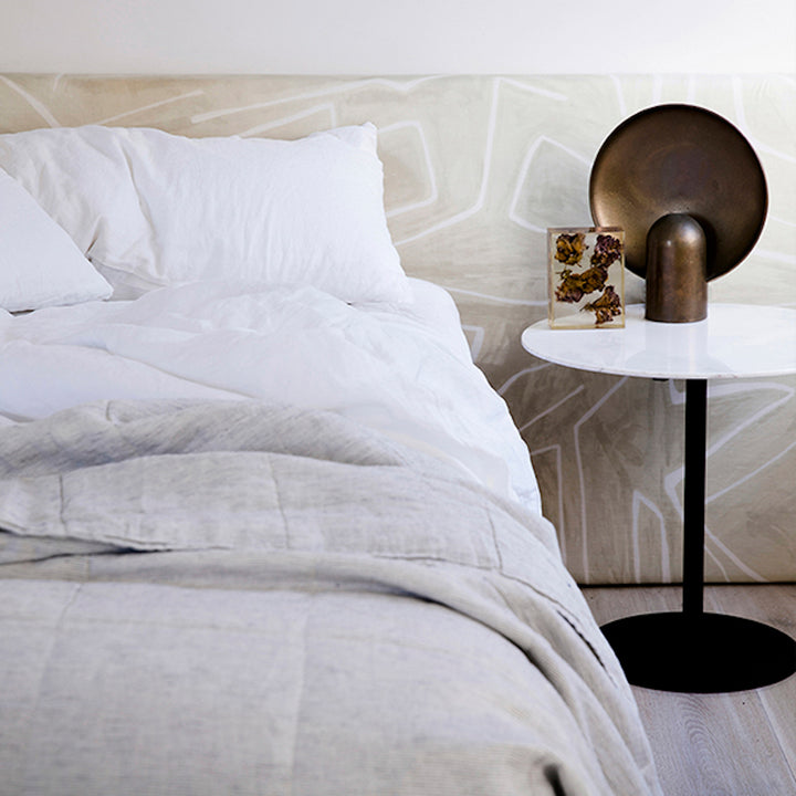 A bed dressed in White bedlinen, with a Pinstripe Quilt Cover, is styled against a printed bedhead and a side table with brass objects. Sizes: Single, Double, Queen, King