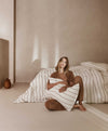 Woman dressed in loungewear sitting on the floor , hugging a pillow next to a bed styled with  cedar stripe linen sheets.