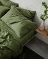 A close up image of a bed styled with forest linen sheet and pillowcases next to a side table with a vase. 