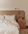 A bedroom styles with a bed dressed in natural linen sheets and pillowcases, next to a  wooden side table.