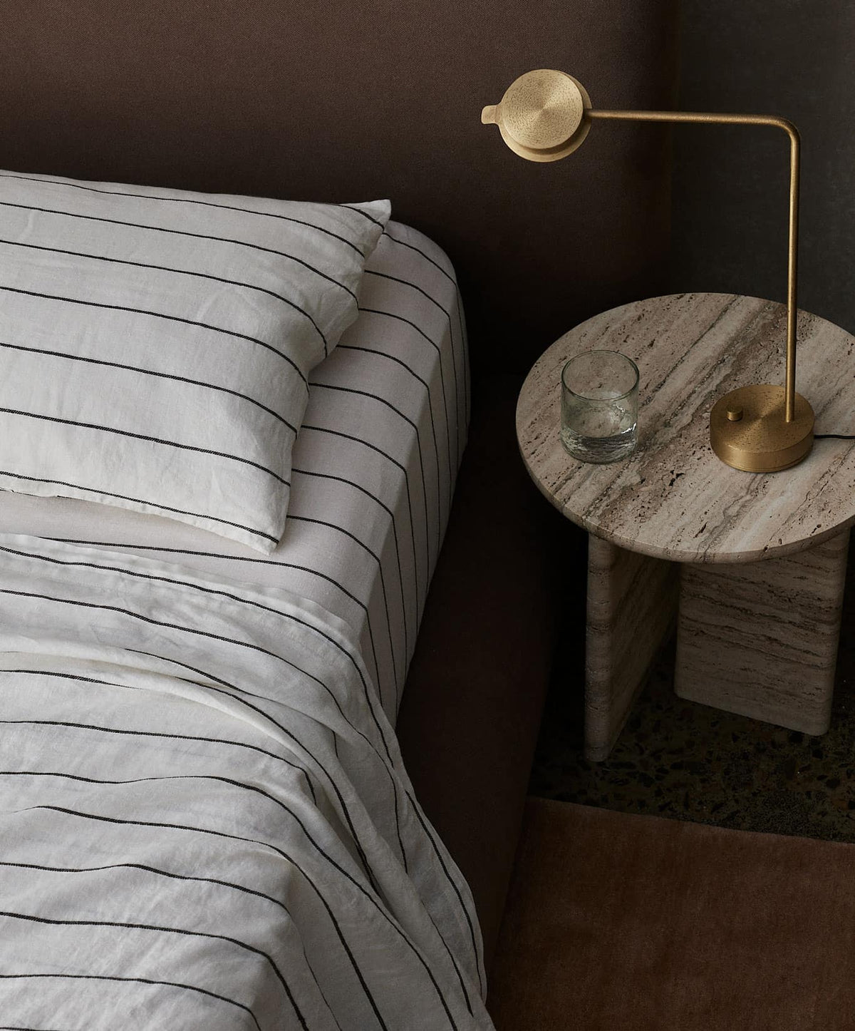 Linen Flat Sheet - Truffle Stripe in natural tones with fine stripes on bed beside round stone table and modern brass lamp