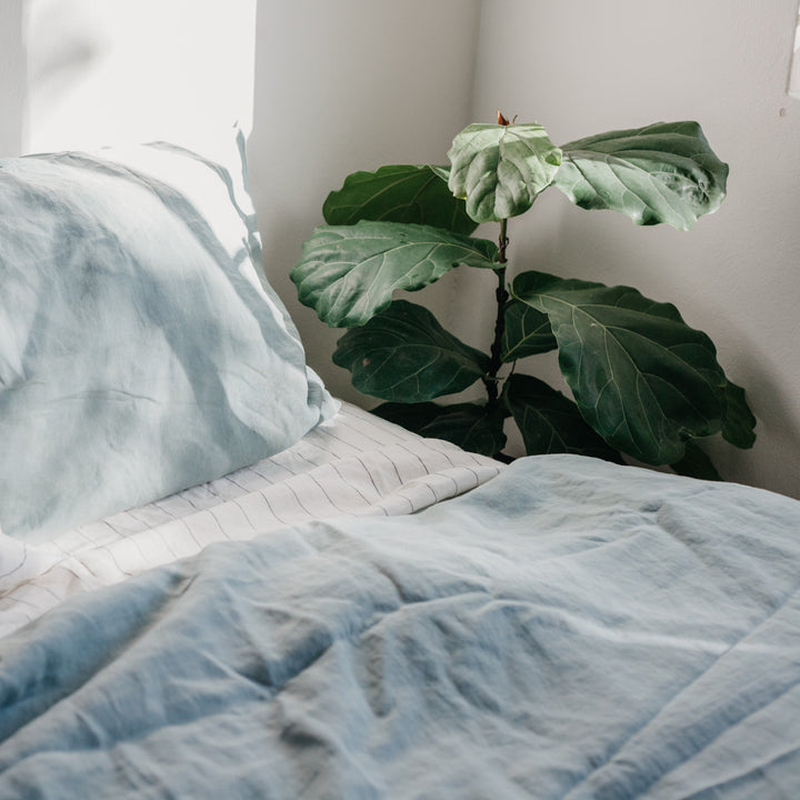 A bed dressed in Sage and Pencil Stripe bedlinen, styled with a green plant.					