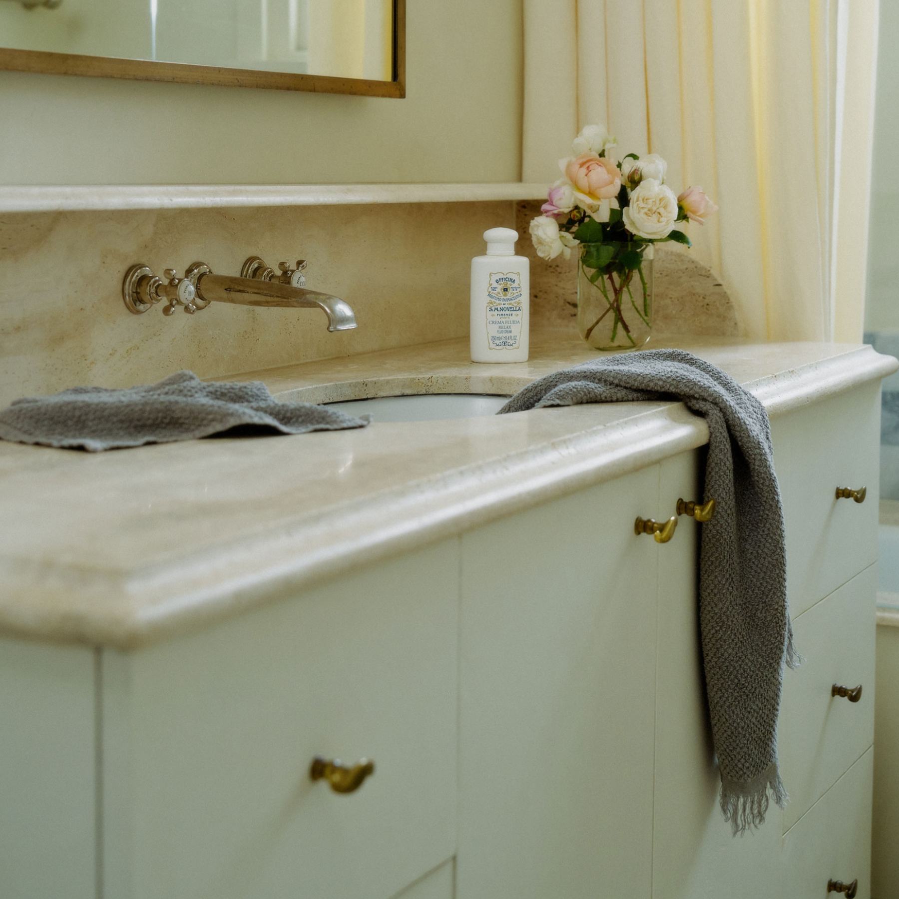 A bathroom vanity with a cream marble countertop, a white pump bottle, a gray towel draped over the edge, and a vase of flowers near a mirror.