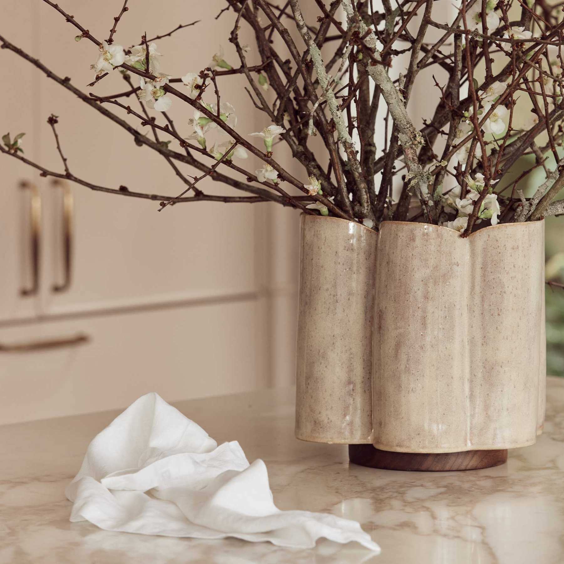 A textured beige ceramic vase with a fluted, three-part cylindrical design, holding dried branches with small white blossoms, set on a light marble countertop with a white cloth nearby.