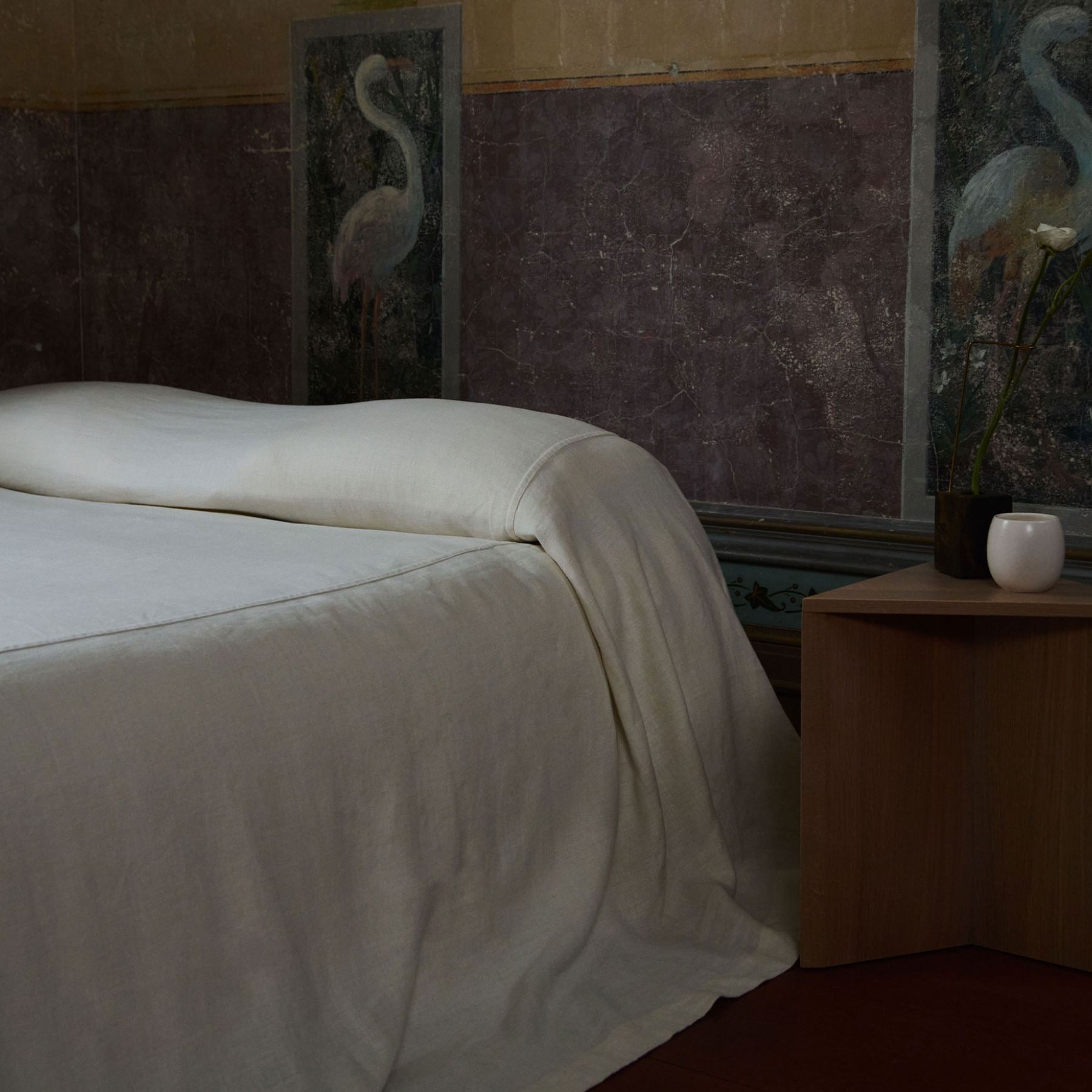 A bed dressed in white bedding, positioned in a dimly lit bedroom with a wooden bedside table, a small white vase, and dark wall panels featuring bird artwork.