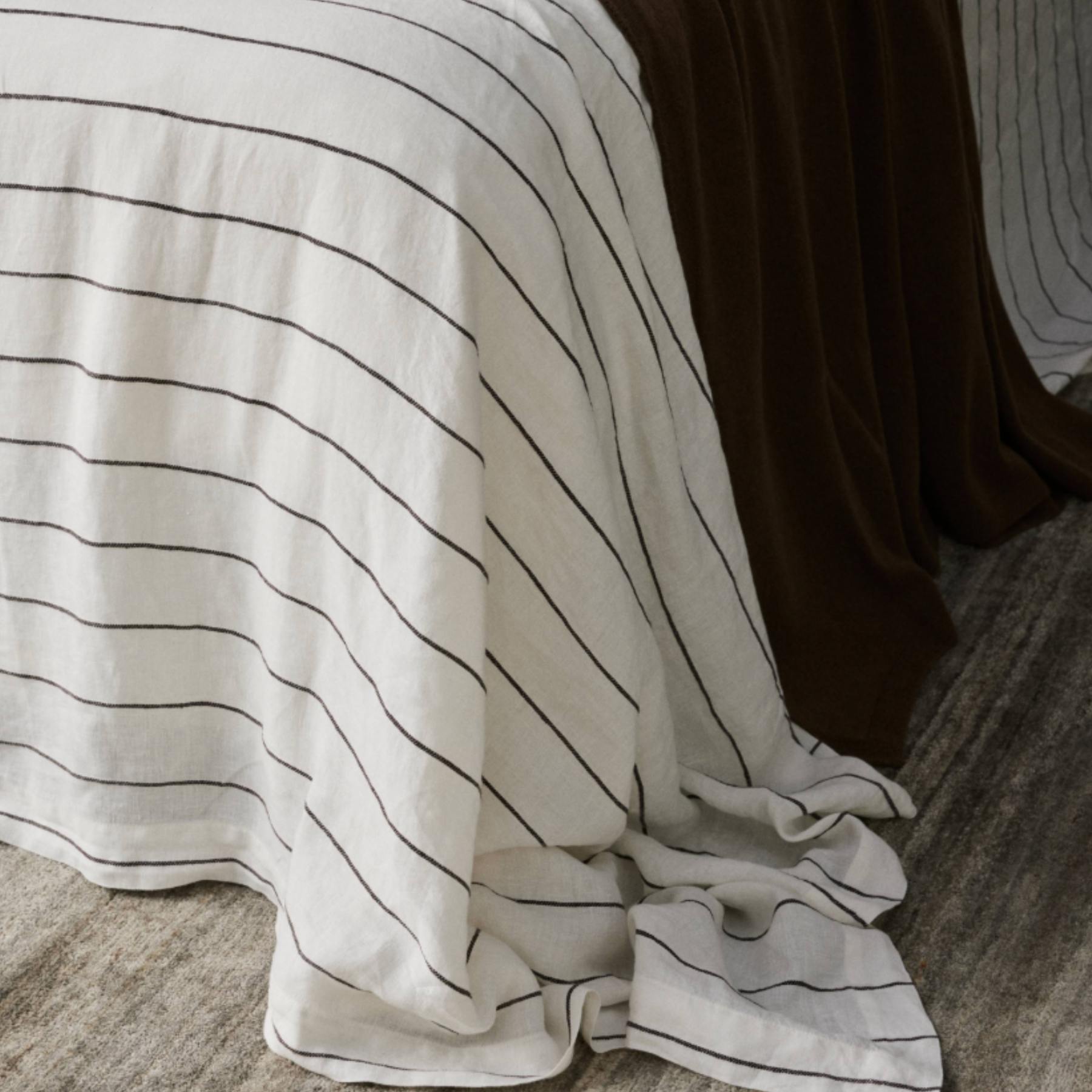 A close-up of a white or off-white fabric textile draped over the edge of a bed, featuring thin black horizontal stripes. A dark brown sheet or duvet is visible behind it, and a textured gray floor is at the bottom.