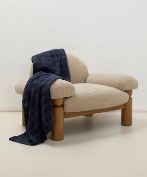 Beige armchair with wooden legs and a navy blanket draped over it against a white wall.
