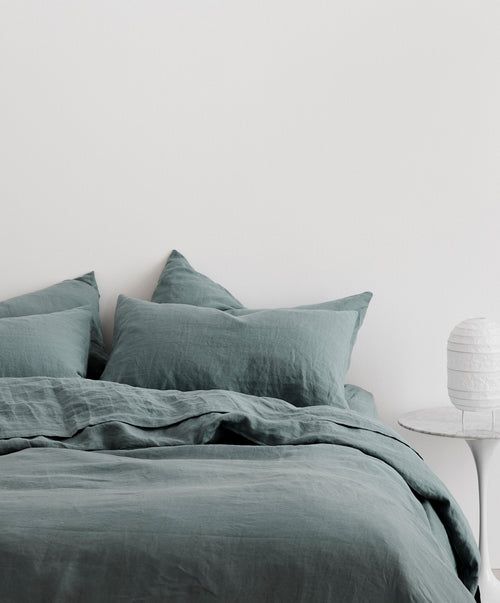 A neatly made bed with the muted blue Linen Duvet Cover - Bluestone and two pillows sits against a white wall. A round white paper lamp on a small side table highlights the elegant duvet cover’s subtle texture.