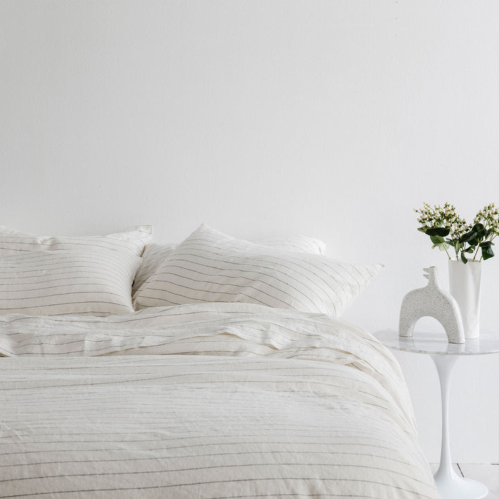 A bed dressed in Pencil Stripe bed linen, Styled with a white bedside table, flowers in a vase and a ceramic ornament. Sizes: Single, Double, Queen, King