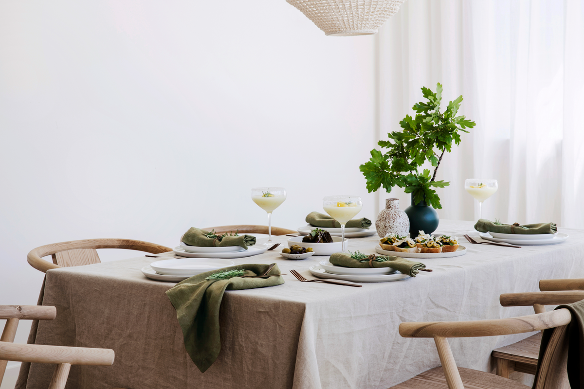 Dining table dressed in natural linen cloth with green napkins, ceramic plates, plants, and soft daylight