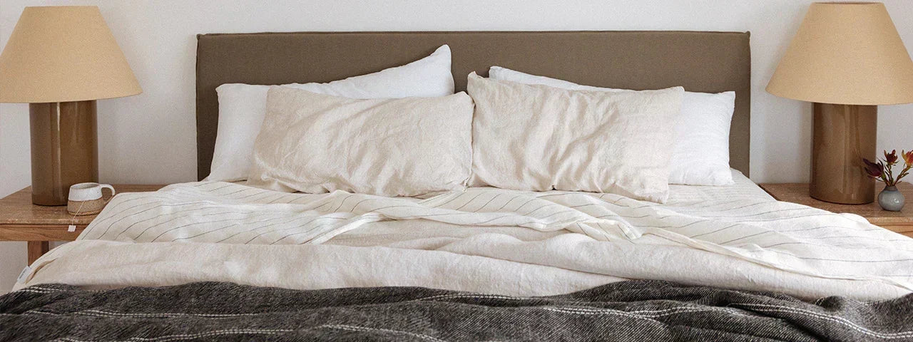 A neatly made bed with off-white bedding, including a duvet cover, multiple pillows, and a dark gray textured throw blanket, set against a brown upholstered headboard. Two matching bedside tables with beige table lamps are visible on either side.