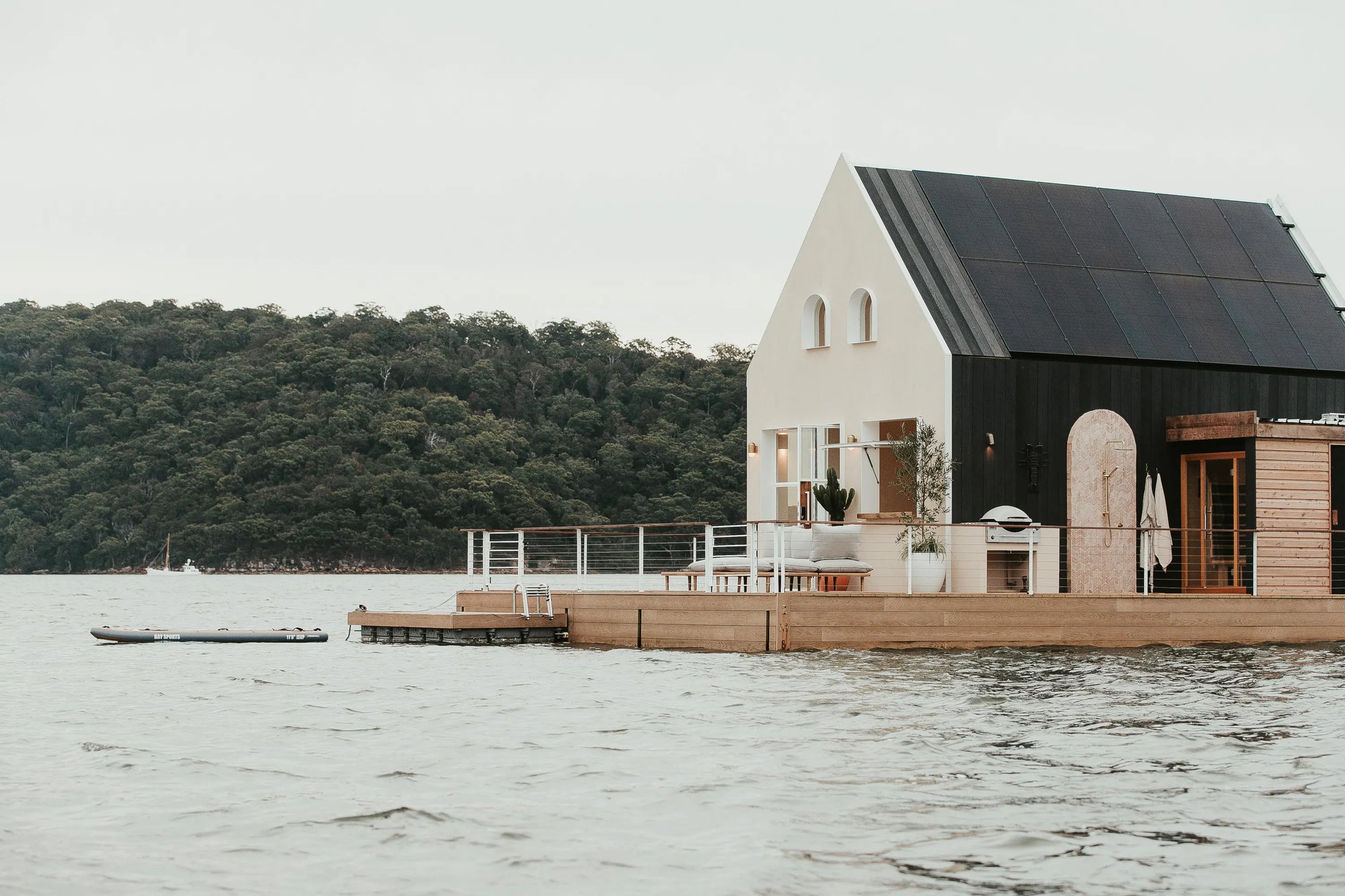 A modern floating house on the water with a white upper section, black lower facade, and wooden deck, set against a backdrop of a tree-covered shoreline under an overcast sky.