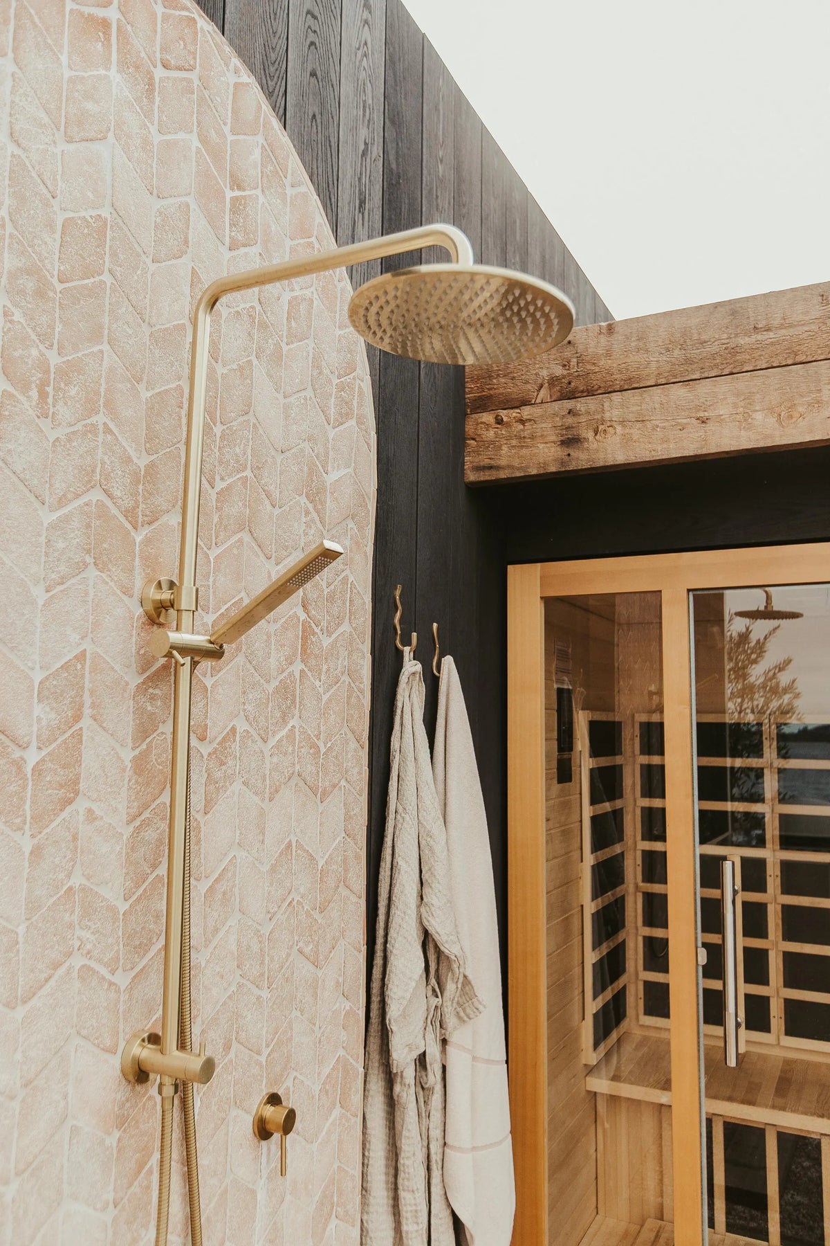 A modern outdoor shower setup with a brushed brass shower head and handpiece mounted against a beige stone wall, next to a dark wood wall and a wooden sauna with hanging towels.