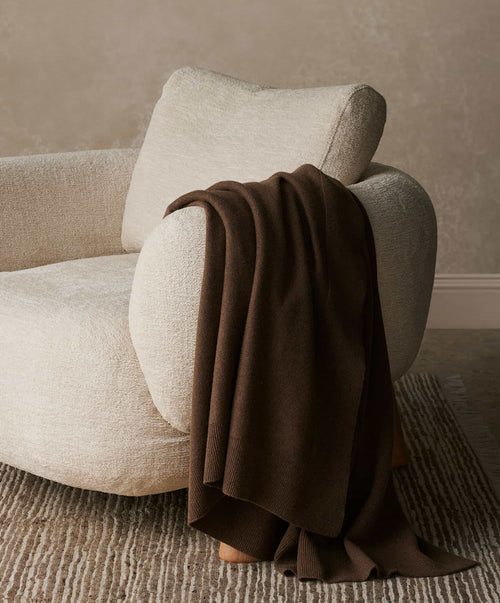 Beige armchair with a brown blanket draped over it against a neutral wall.
