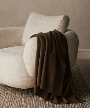 Beige armchair with a brown blanket draped over it against a neutral wall.