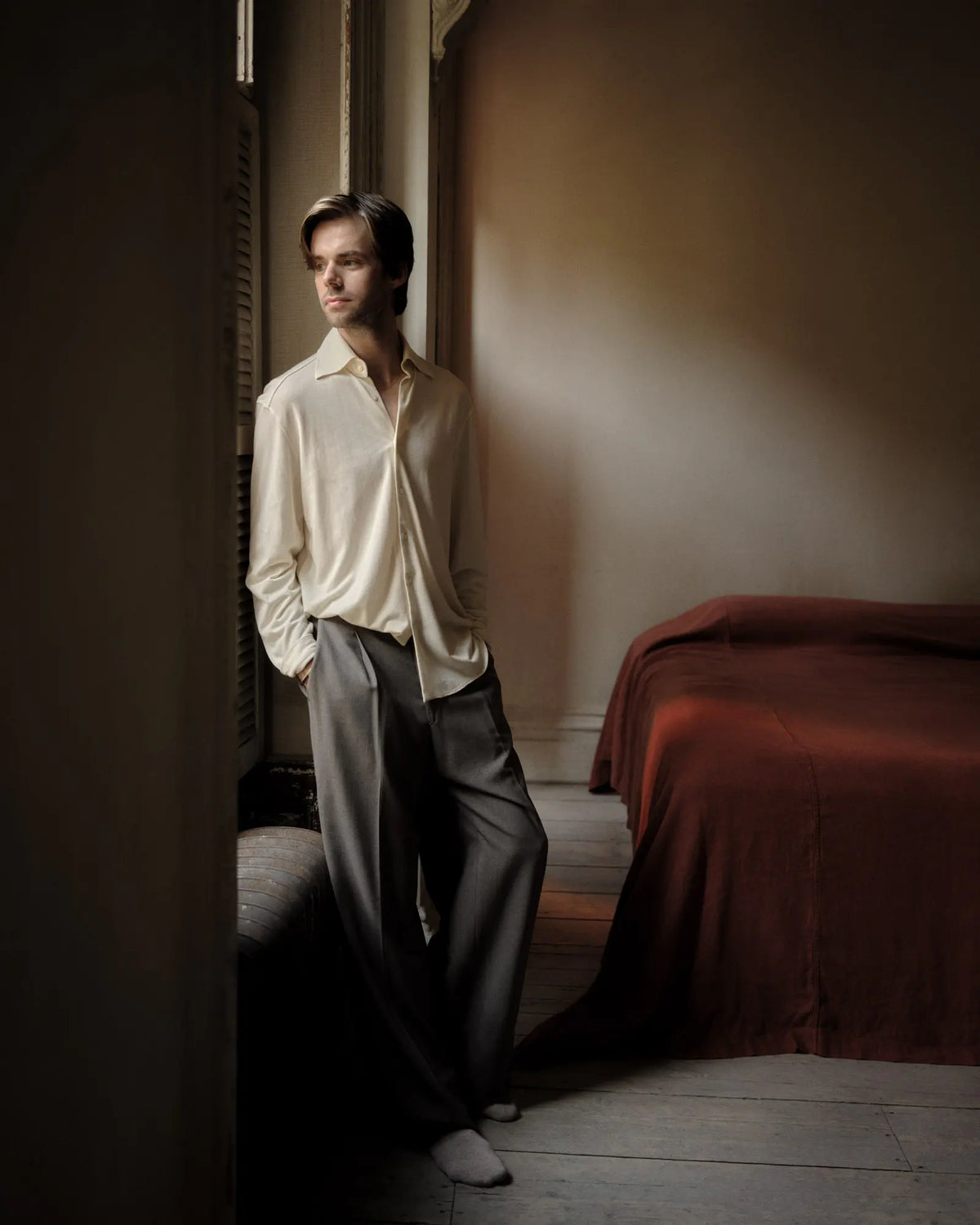 Man in a white shirt and gray pants standing in a dimly lit room with a bed.