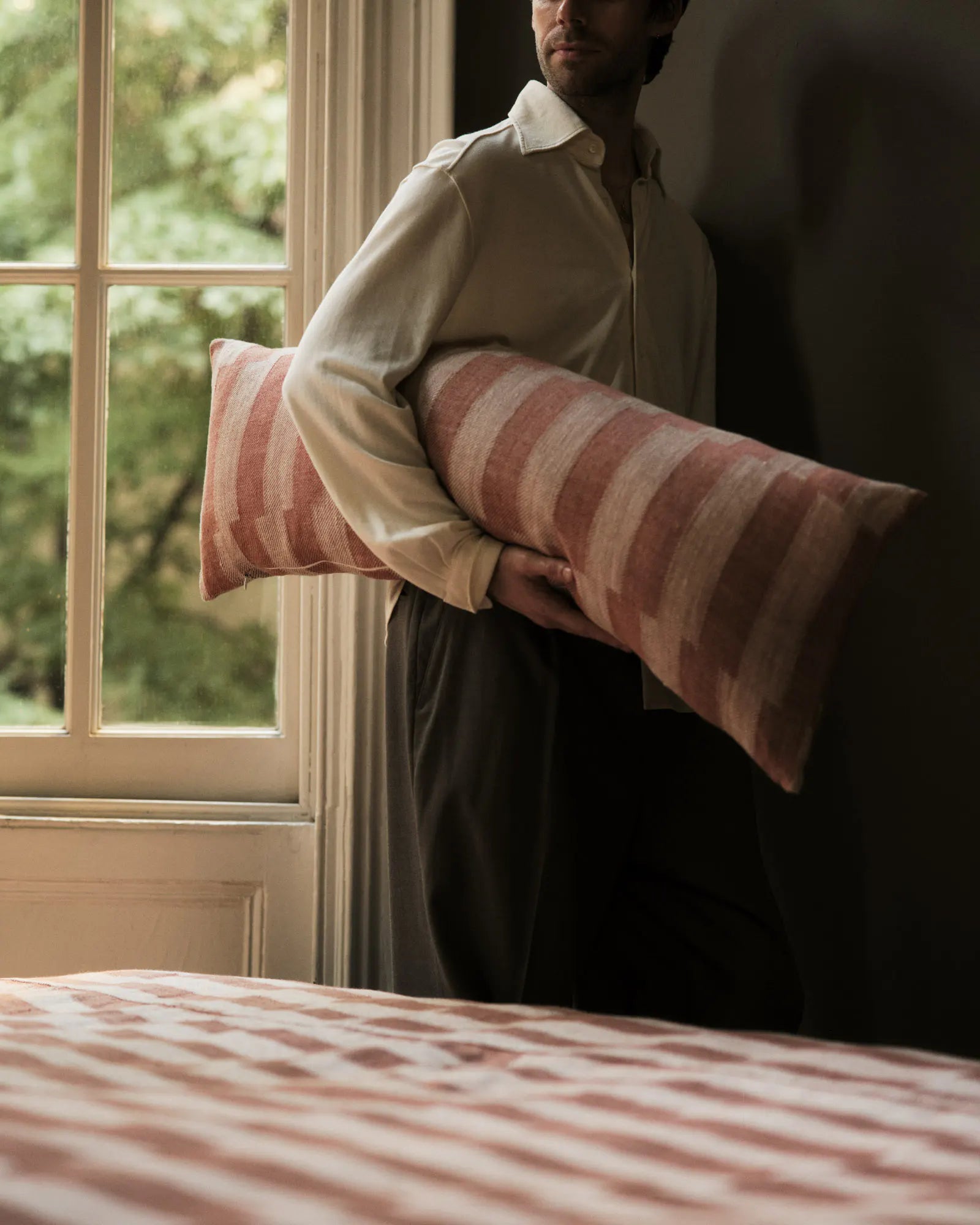 A man in a cream shirt and dark trousers holds a striped lumbar pillow with pink and beige stripes next to a window. The matching striped bedding is visible in the foreground.