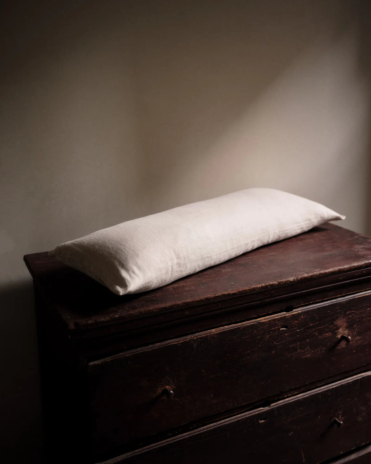 A rectangular off-white pillow resting on a dark brown wooden chest of drawers against a warm beige wall.