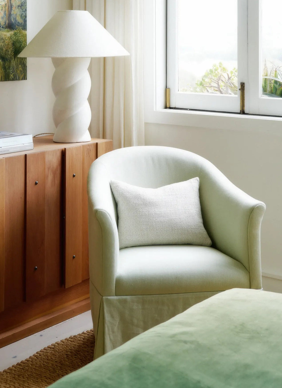 Softly lit corner with a curved armchair, textured cushion, sculptural lamp, and sheer white curtains by a window