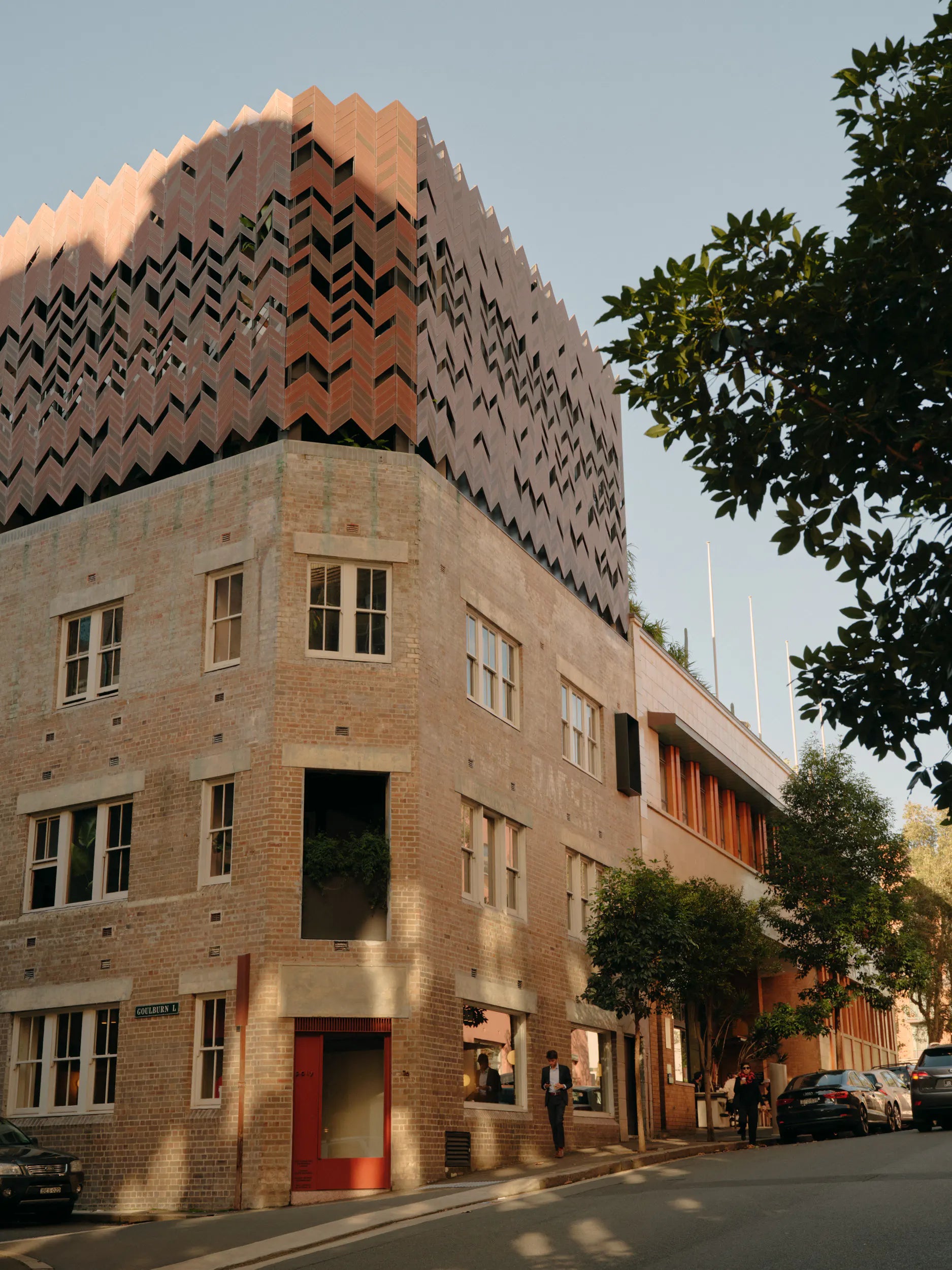 A corner brick building with a distinctive patterned terracotta upper facade, shown on a city street with trees, parked cars, and a few people walking nearby.