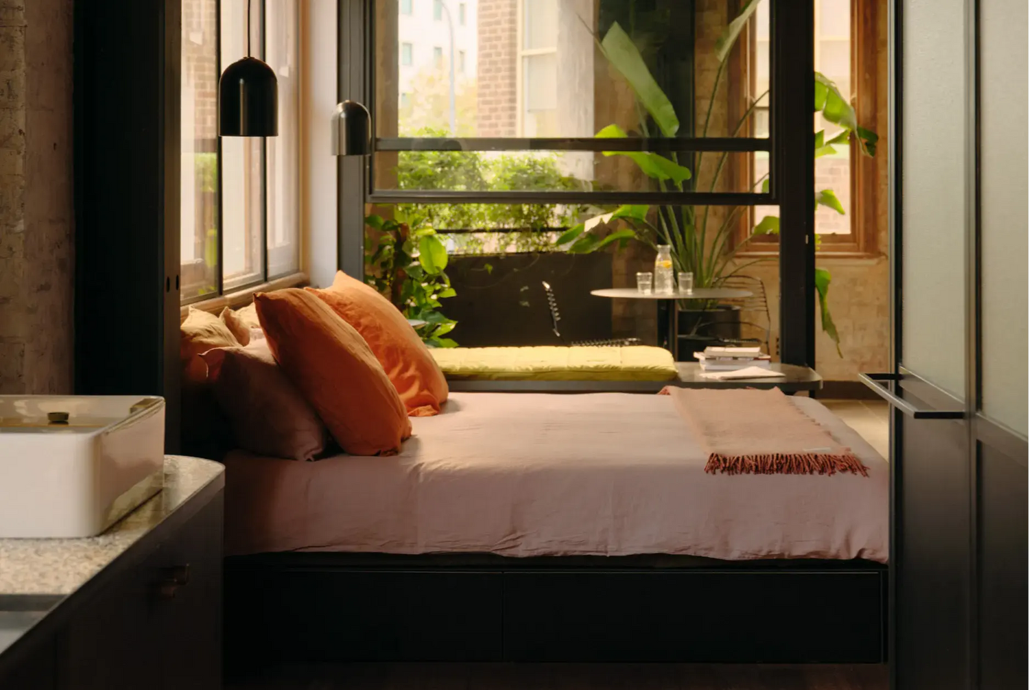 A modern bedroom interior featuring a low black platform bed with blush pink bedding, orange and brown pillows, and a small orange throw blanket. The bed is positioned in front of a large black-framed glass window looking out onto a green outdoor patio.